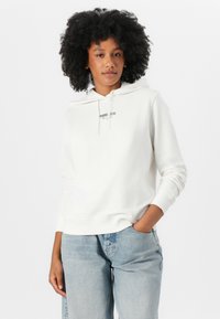 Witte hoodie van zacht materiaal, met een kangoeroezak, verstelbare trekkoordcapuchon en een bedrukte logo op de borst. Gepaarde met lichtblauwe jeans.