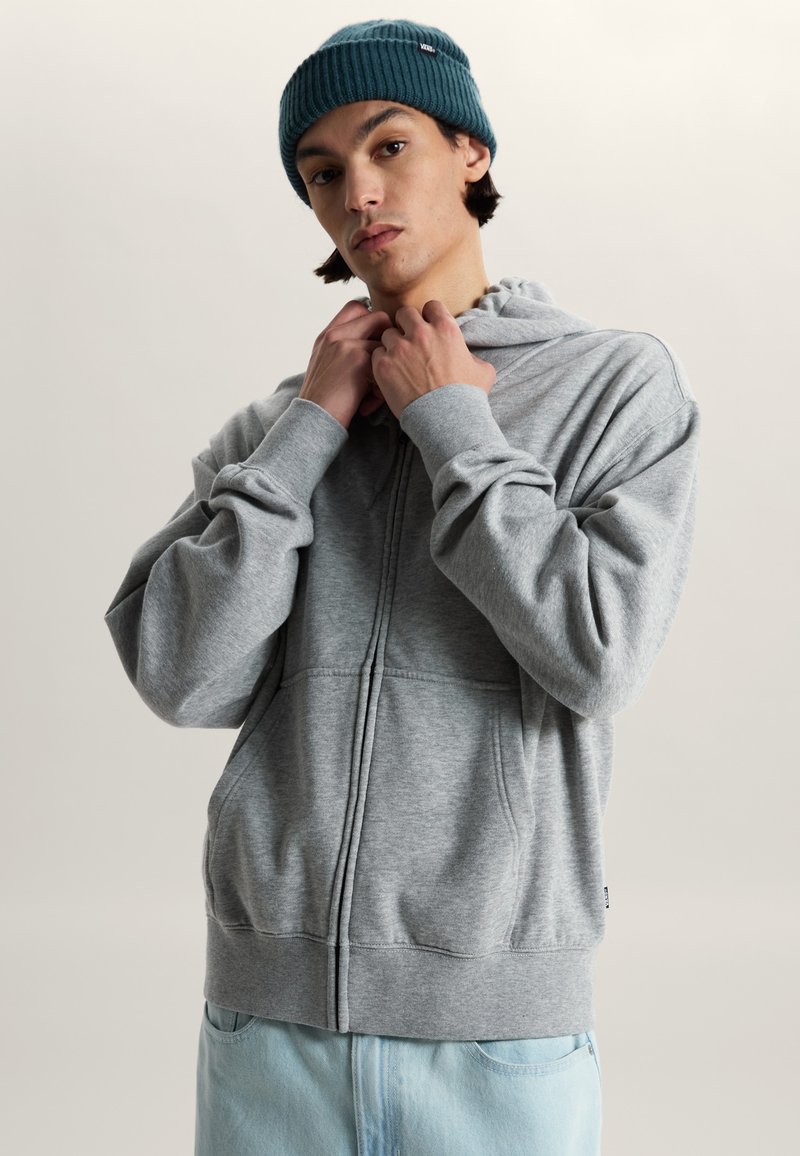 Junger Mann, der einen grau zu knöpfenden Hoodie anpasst, trägt eine dunkelgrüne Mütze und hellblaue Jeans vor einem einfarbigen Hintergrund.