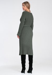 Strickkleid in Dunkelgrün mit geripptem Textur, langen Ärmeln und Gürtel-Detail in der Taille, kombiniert mit hohen schwarzen Wildleder-Stiefeln mit Absatz.