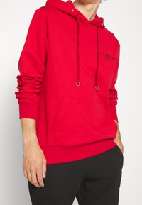 Sudadera roja con capucha, cordones y un bolsillo frontal. Presenta una impresión de logo en el pecho. Hecha de una tela suave y texturizada.