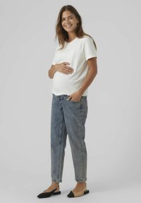 T-shirt bianco in cotone, vestibilità comoda, abbinata a jeans azzurri chiari. La modella indossa scarpe basse nere e beige, in piedi con le mani sulla pancia.