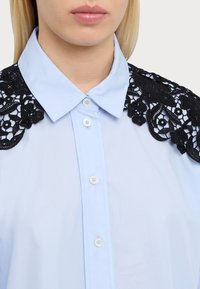 Cămașă bleu deschis cu un guler din dantelă neagră cu modele florale, închidere cu butoane în față și un material fin, ușor.