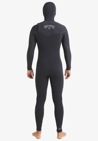 Osoba ubrana w czarny, jednoczęściowy piankowy skafander z kapturem, stojąca boso i odwrócona plecami, z widocznym logo "Billabong" na górnej części pleców i dolnej części nogi.