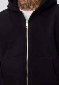 Homme portant un sweat à capuche noir zippé sur un t-shirt blanc, montrant un tatouage floral sur le cou et des détails de fermeture éclair argentée.