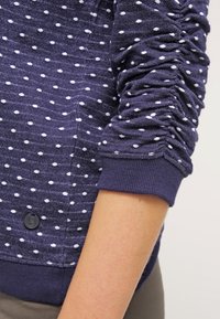 Morska bluza z niebieskimi rękawami w białe kropki, marszczony materiał, ściągacz na mankietach oraz guzik przy dolnym wykończeniu, noszona na ramieniu osoby.