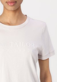 Lys grå kortærmet t-shirt lavet af bomuld, med rund halsudskæring og diskret broderet branding på brystet. Glat tekstur.