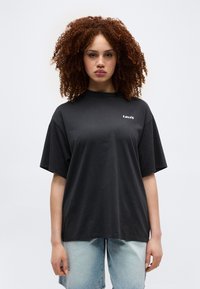 Γυναίκα με σγουρά κόκκινα μαλλιά, φορώντας ένα μαύρο υπερμεγέθες μπλουζάκι Levi's και ανοιχτόχρωμα μπλε τζιν σορτς, στέκεται μπροστά σε μια απλή ανοιχτόχρωμη επιφάνεια.