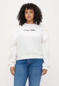 Hvid sweatshirt med rund halsudskæring, lange ærmer, broderet "Tommy Hilfiger" logo og guld dekorative knapper på skulderen. Sammen med blå jeans.