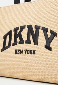 Sac fourre-tout en paille tannée avec le logo brodé en noir "DKNY NEW YORK", surface texturée et poignées noires solides.
