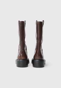 Botas de cuero marrón hasta la rodilla con punta redondeada, suela de goma negra resistente y tiradores en la parte superior para ponerlas y quitarlas fácilmente.