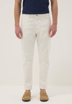 Hombre vestido con pantalones blancos de corte ajustado, camisa azul marino y mocasines de ante color tan, de pie contra un fondo claro y sencillo.