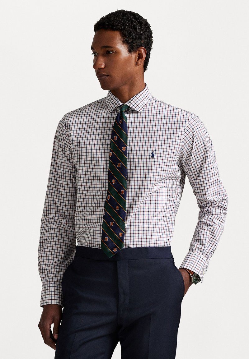 Camisa blanca de botones con un patrón de cuadros en rojo y azul marino, acompañada de una corbata verde oscura con rayas diagonales y pequeños emblemas.