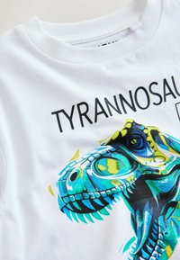 Vit bomullströja med en livlig blå och gul Tyrannosaurus rex-grafik med svart text. Mjuk textur och rundad halsdesign.