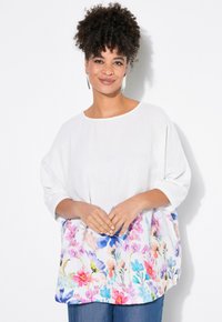 Top bianco con tessuto texturizzato e maniche ampie, che presenta una stampa floreale colorata sulla parte inferiore. Abbinato a jeans blu.
