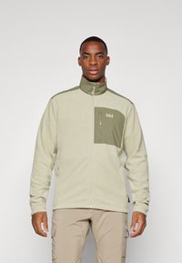 Chaqueta de forro polar beige claro con acentos verde oliva. Presenta un cuello alto y un bolsillo en el pecho con cremallera y un logo. Textura suave, ajuste relajado.