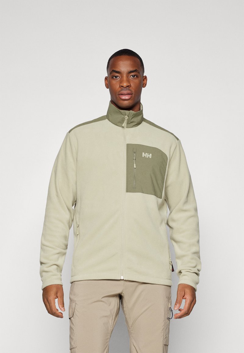 Chaqueta de forro polar beige claro con acentos verde oliva. Presenta un cuello alto y un bolsillo en el pecho con cremallera y un logo. Textura suave, ajuste relajado.