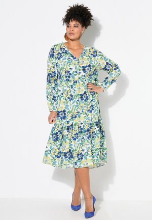 Blumenmusterkleid in Grün-, Gelb- und Blautönen, mit langen Ärmeln, Knopfdetails und einem gestuften Rock. Kombiniert mit blauen Schuhen.