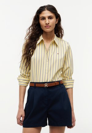 Frau mit langen lockigen Haaren, die ein gelb gestreiftes Hemd mit Knöpfen, marineblaue Shorts und einen braunen Gürtel mit goldener Schnalle trägt.