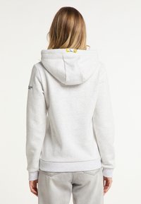 Žena s dlouhými vlasy oblečená v světle šedé mikině s žlutými očky na kapuci, v kombinaci se světle šedými džíny, zobrazená zezadu.