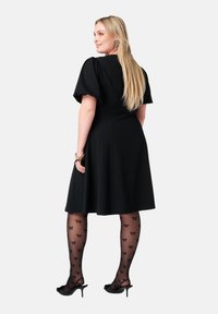 Zwarte jurk met korte pofmouwen en een uitlopende rok. Draagt zwarte panty’s met kleine strikmotieven en hakken.