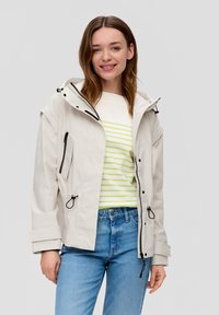 Junge Frau lächelt, trägt eine hellbeige Kapuzenjacke, ein gestreiftes weißes und grünes T-Shirt und eine blaue Jeans, steht vor einem einfarbigen Hintergrund.