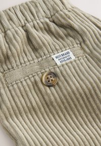 Tissu en velours côtelé beige avec texture côtelée, comprenant une poche arrière et un bouton rond. L'étiquette indique "Next Brand Authentic Est. 1982."