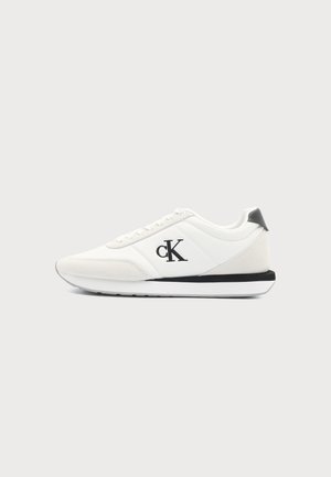 Basket basse blanche avec logo noir "cK" sur le côté et languette de talon noire, dotée de lacets blancs et d’une semelle avec des accents noirs.