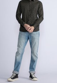 Camicia a maniche lunghe a fantasia scura abbinata a jeans azzurri chiari, con le maniche arrotolate e sneakers nere alte con suole bianche.