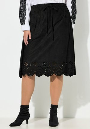 Černá kolenní sukně s květinovým laserem řezaným lemem a stahovacím pasem, nošená s bílou blůzou a černými kotníkovými botami na podpatku.
