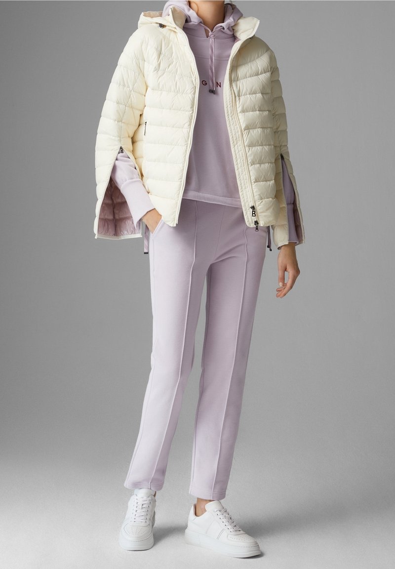 Kräppjacka i krämfärg med kviltad design, tillsammans med en lila huvtröja och matchande joggers, avslutad med vita sneakers.