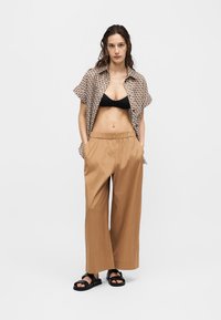 Γυναίκα στέκεται με τα χέρια στις τσέπες φορώντας κοντομάνικο πουκάμισο με σχέδιο ανοιχτό πάνω από μαύρο bralette, μπεζ παντελόνι με φαρδιά πόδια και μαύρα σανδάλια.