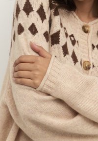 Beige stickad cardigan med en diamantmönster i mörkbrunt, ribbade ärmslut och stora runda knappar, med texturerad tyg och en avslappnad passform.