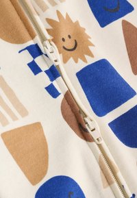 Tela suave multicolor con formas geométricas y patrones de soles sonrientes. Cierre central con un tirador de plástico sobre un fondo crema.