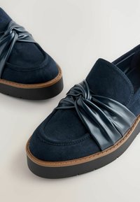 Navy blue suede loafers with a knotted glossy leather strap, thick rubber sole, and contrasting brown accent on the base.