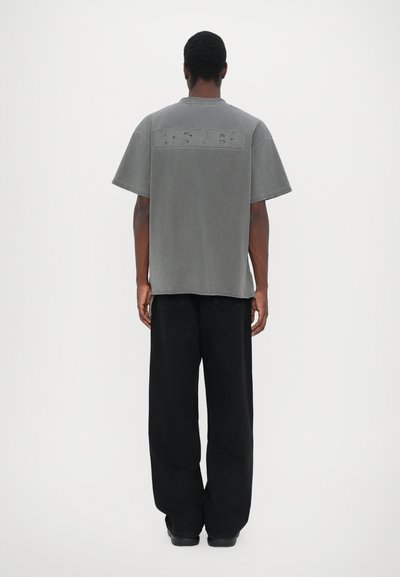 T-shirt oversize en coton gris à manches courtes avec logo "KSUBI" en relief dans le dos, associé à un pantalon noir ample.