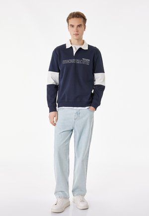 Jonge man staand, gekleed in een donkerblauwe trui met witte kraag en strepen, lichtblauwe jeans en witte sneakers, tegen een witte achtergrond.