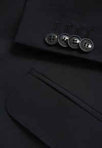 Czarna tkanina garniturowa cechuje się gładką teksturą, kątową kieszenią oraz czterema okrągłymi czarnymi guzikami z czterema otworami każdy. Widoczne schludne przeszycia.