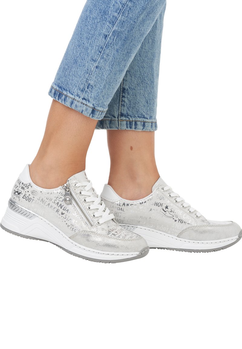 Baskets blanches avec des accents métalliques, présentant un dessus texturé avec des mots imprimés, un laçage à l'avant et une semelle blanche épaisse.