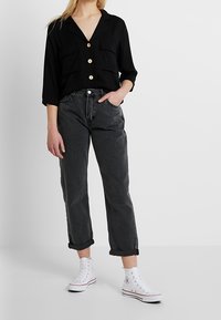 Femme portant une chemise noire boutonnée, un jean gris foncé retroussé et des baskets blanches montantes, debout avec une main dans la poche.