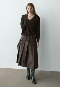 Brauner gestrickter V-Ausschnitt-Pullover, kombiniert mit einem braunen plissierten Midirock und kniehohen schwarzen Lederstiefeln. Strukturierter Stoff mit dezenten Akzenten.