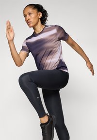 Kortermet treningstopp med et lilla og brunt gradientmønster, kombinert med sorte tights og svarte treningssko, på en person i bevegelse.