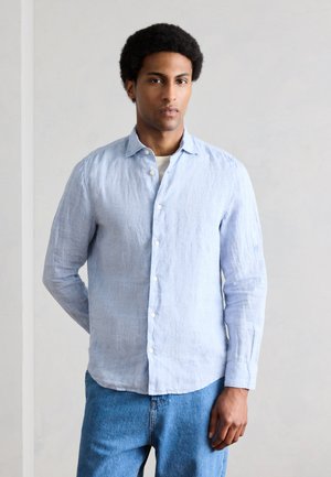 Ung mand med kort krøllet hår iført en lyseblå skjorte med knapper over en hvid undertrøje og blå jeans, stående mod en ensfarvet baggrund.