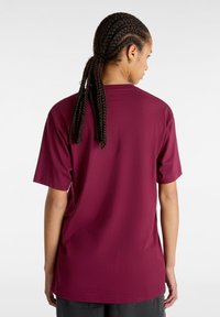 Personne aux longs cheveux tressés attachés en arrière, vêtue d'un t-shirt uni rouge foncé à manches courtes, vue de dos sur fond blanc.
