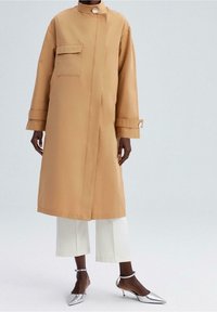 Długi, beżowy płaszcz trench z wysokim kołnierzem, przednią kieszonką i srebrnymi elementami. W zestawieniu z białymi spodniami 7/8 i srebrnymi szpilkami.