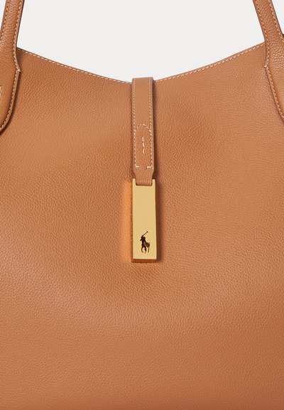 Tan læderhåndtaske med tekstureret overflade, prydet med et hardware-tag i guld med logo. Tasken har en bred åbning og forstærkede håndtag.