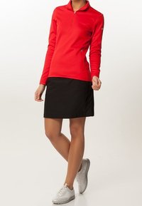 Mujer vestida con una camiseta roja de manga larga con cremallera, falda negra y zapatillas blancas, de pie con las piernas cruzadas contra un fondo liso.