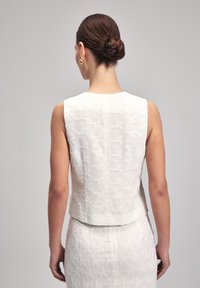 White sleeveless top with textured floral embroidery, featuring a rounded neckline and a fitted silhouette, seen from the back.