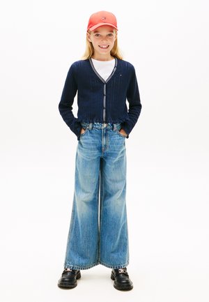 Χαμογελαστή νεαρή κοπέλα με κόκκινο καπέλο, μπλε ζακέτα, λευκό πουκάμισο, μπλε τζιν παντελόνι με φαρδιά πόδια και μαύρα παπούτσια, με τα χέρια στις τσέπες, σε λευκό φόντο.