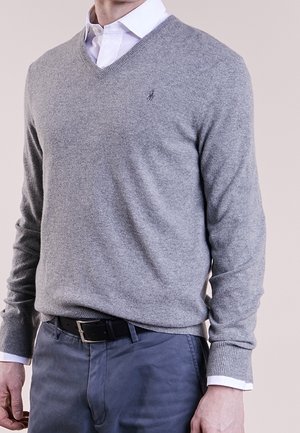 Mężczyzna ubrany w szary sweter w serek na biały koszulę z kołnierzykiem, niebieskie spodnie i czarny pasek, stojący na jednolitym tle.