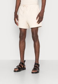 Personne à la peau foncée portant un short à cordon crème et des sandales en cuir noir, debout devant un fond blanc.